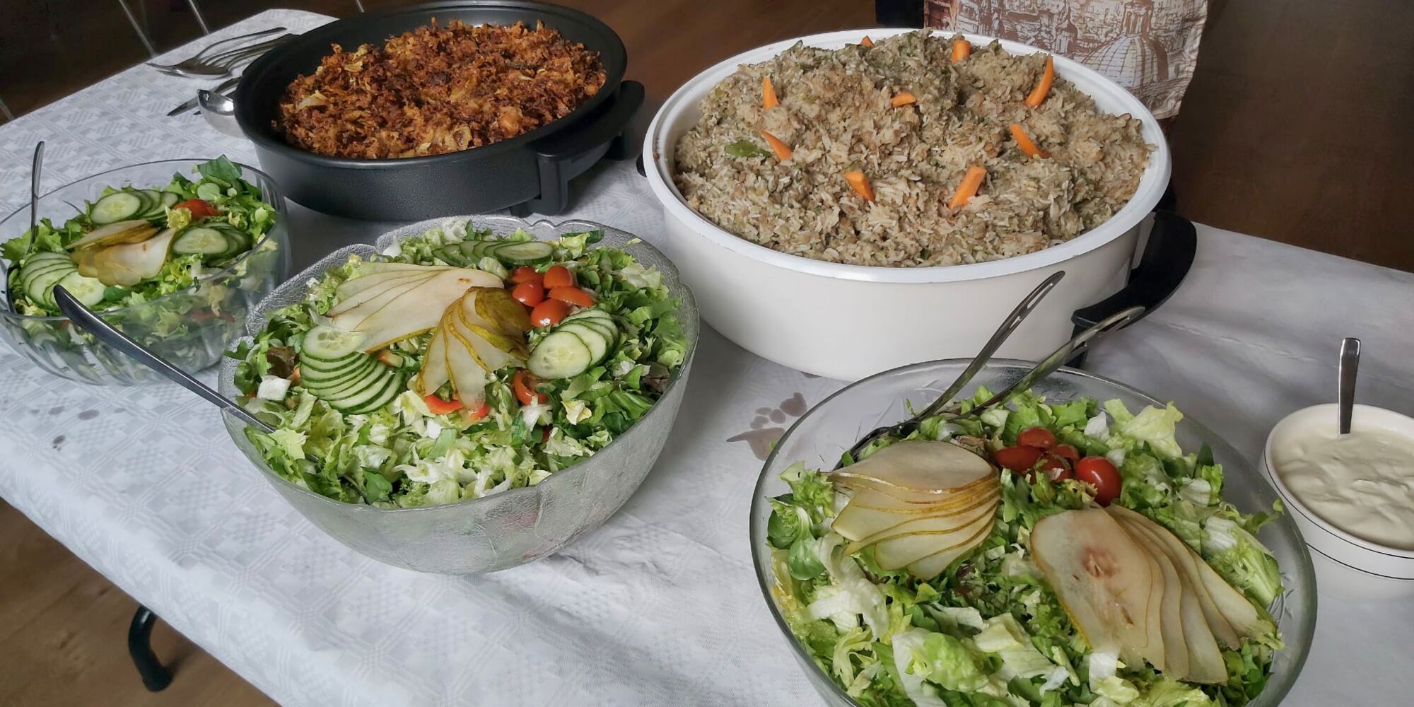 Hier sieht man mehrere Speisen in Schüsseln und Warmhaltepfannen, unter anderem vorne drei Salate.