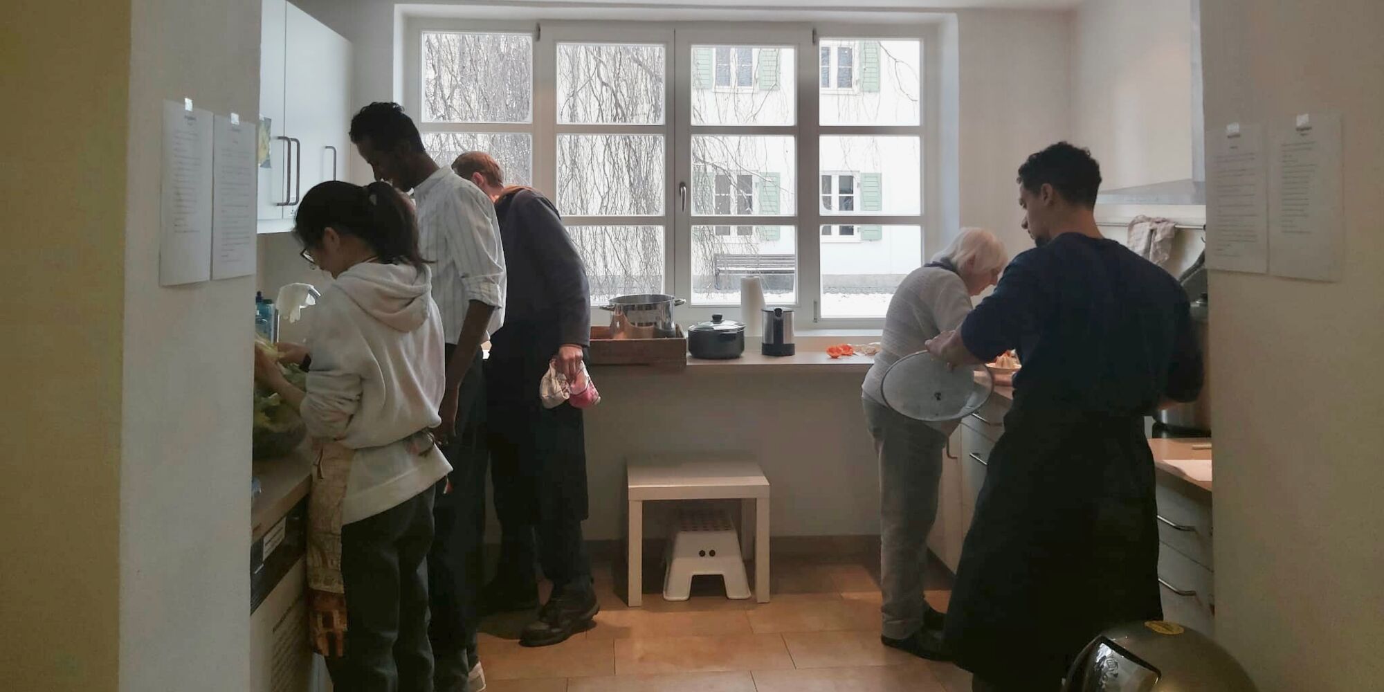 Hier sieht man mehrere Menschen, die links und rechts jeweils an Küchenzeilen stehen und kochen oder abspülen.