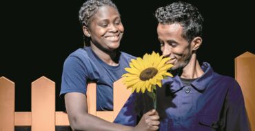 Hier sieht man links eine junge Frau, die einem jungen Mann rechts lächelnd eine Sonnenblume über einen Zaun reicht. Der junge Mann sieht die Blume an. Beide tragen dunkelblau und sind schwarz.