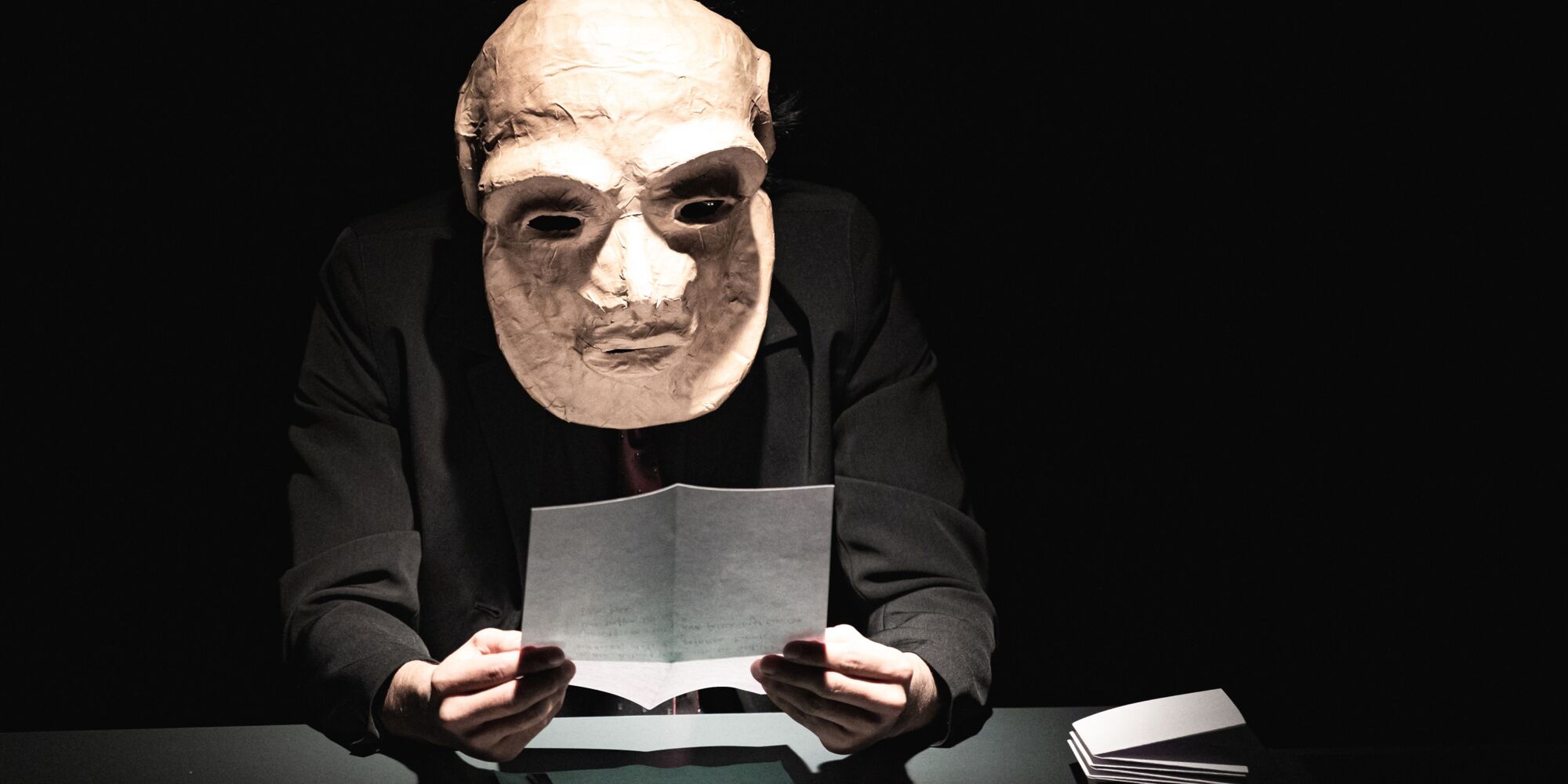 Hier sieht man einen der Darsteller unseres Benefizabends 2022. Er sitzt, trägt eine große Maske und liest einen Brief.