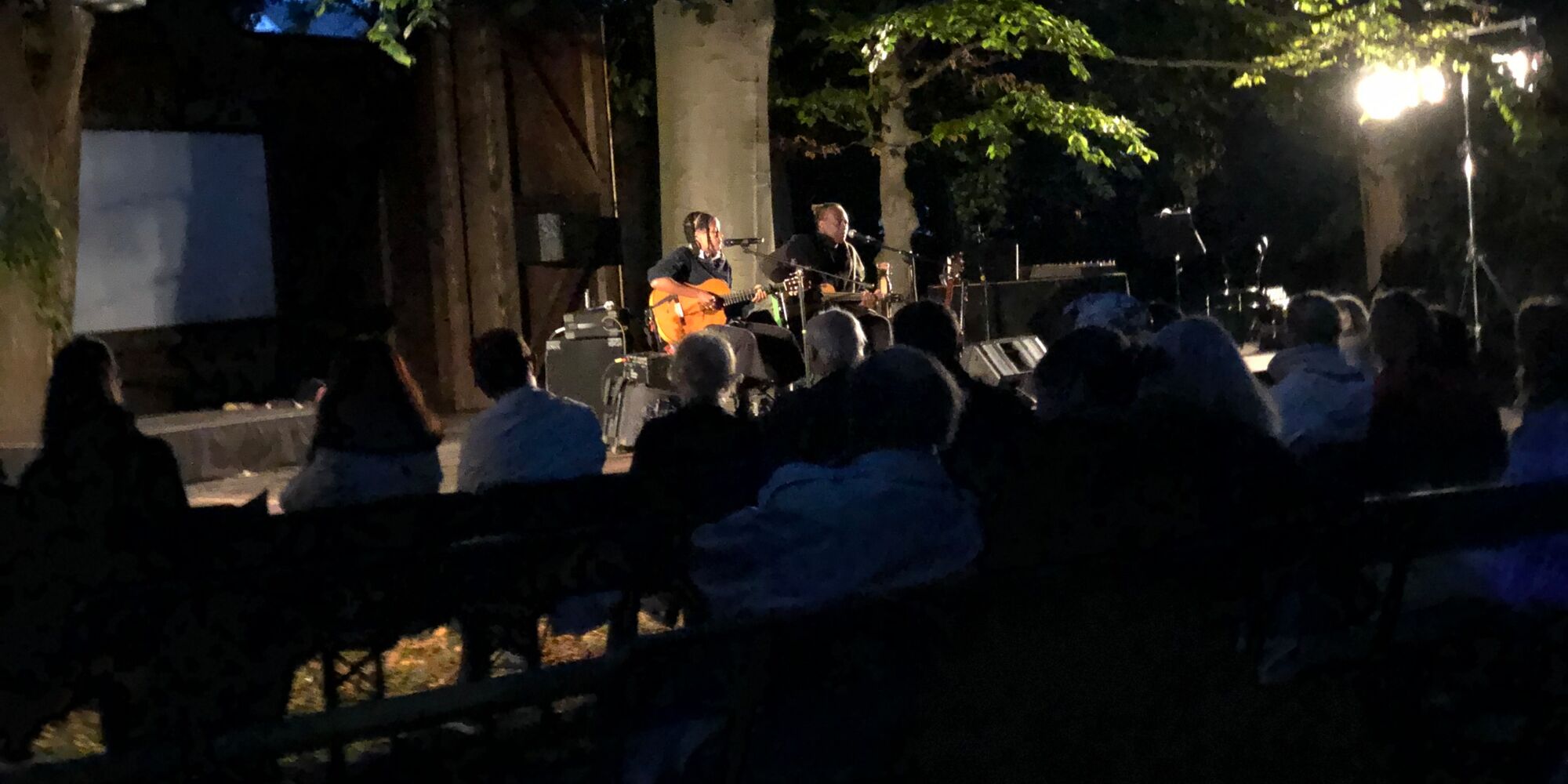 Hier sieht man Wally und Ami Warning auf der Freilichtbühne der Mohr-Villa Freimann am Abend. Sie haben jeweils eine Gitarre und ein Mikrofon vor sich. Im Vordergrund ist das Publikum zu erkennen. Auf der Bühne stehen zwei große Bäume.