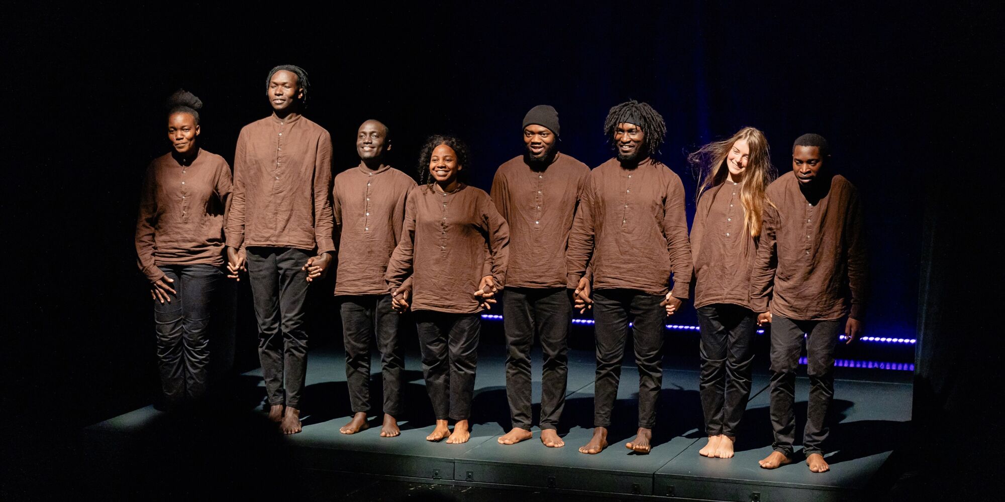 Hier sieht man die acht Darstellenden der Produktion "Balance" beim Schlussapplaus. Sie halten die Hände und tragen alle das gleiche Kostüm bestehend aus einer schwarzen Hose und einem braunen lockeren Hemd.