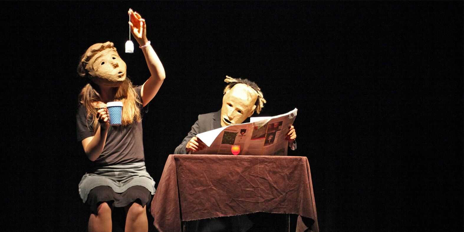 Dies ist ein Szenenfoto aus "Unterm Sprung". Zwei Schauspieler sitzen auf der Bühne und tragen Masken. Die ganz links hält eine Teetasse in der einen und einen Beutel in der anderen Hand den sie ansieht. Der andere sitzt am Tisch und liest Zeitung.