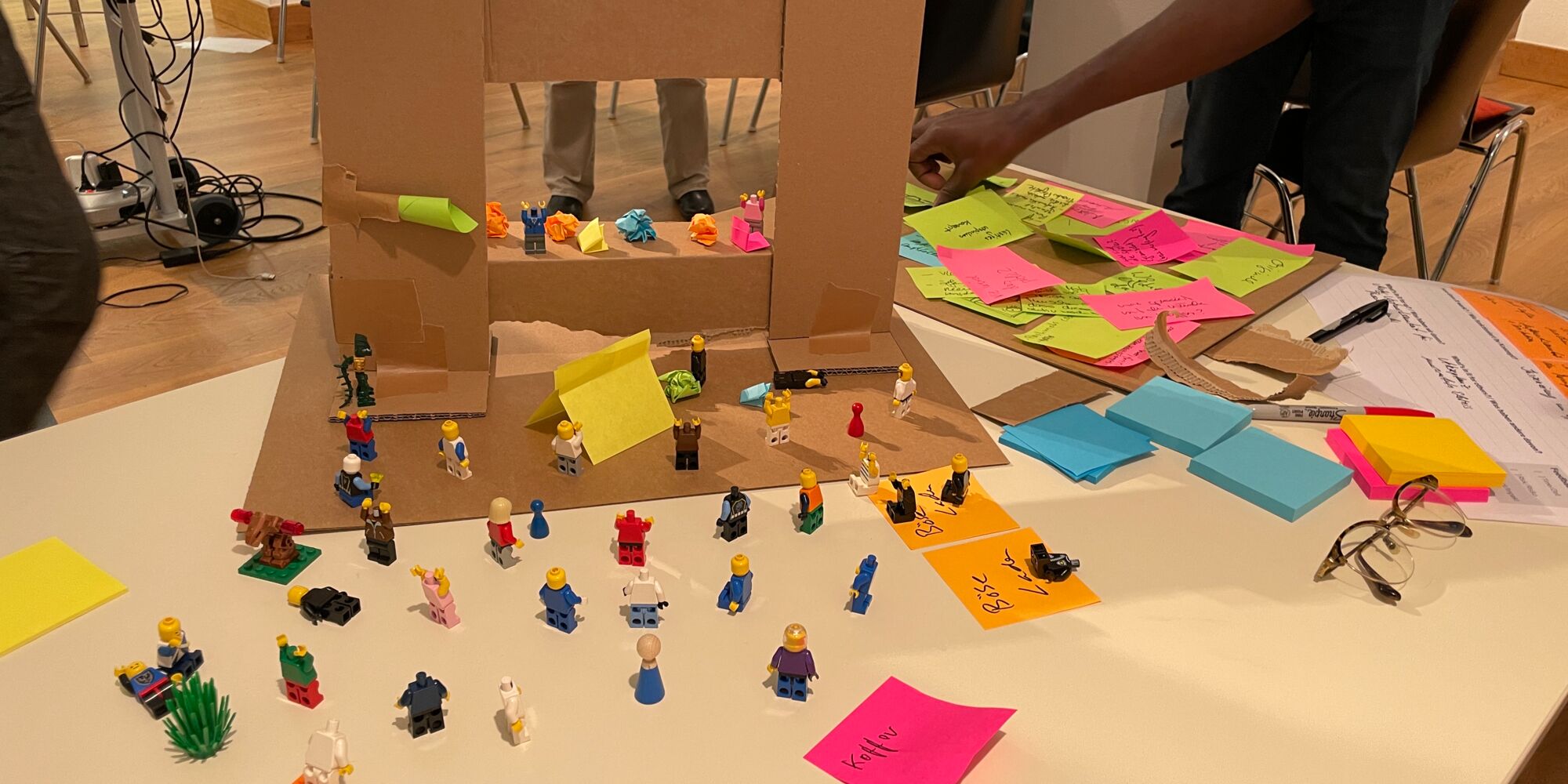 Hier sieht man ein Modell aus Pappe und vielen Legofiguren. Im Hintergrund stehen Menschen um das Modell, ein Arm zeigt von der Seite auf das Modell.