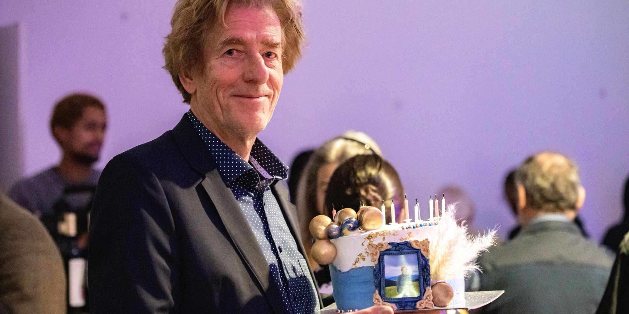 Hier sieht man man Viktor Schenkel auf unserem Benefizabend 2022. Er hält eine reich dekorierte Torte in der Hand, die er als nachträgliches Geburtstagsgeschenk erhalten hat. Er lächelt in die Kamera.