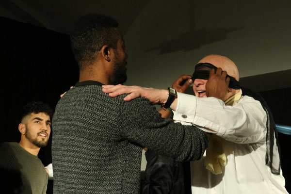 Hier sieht man ein Foto aus dem Stück "Orient Connection". Ein älterer Mann hat eine Augenbinde um die ihm gerade von einem jüngeren Mann abgenommen wird. Der ältere hat seine Hände auf den Schultern des jüngeren Mannes.